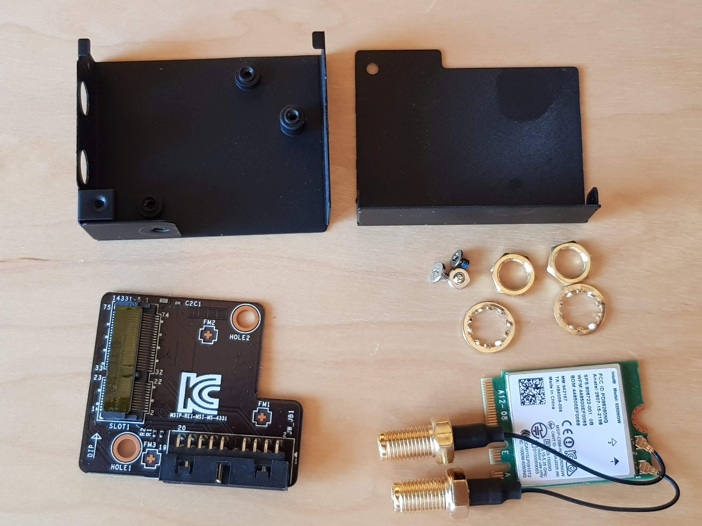 The whole WiFi/Bluetooth module disassembled. On the top of the photo there are two parts of the metal cover. On the bottom left side of the image there's the adapter – it has only the connectors and two resistors on it. The M.2 connector is missing – per description on the silk mask – pins from 24 to 31. On the right bottom side of the image there's a set of screws and the WiFi/Bluetooth card itself with two anntena connectors attached. It has an M.2 connector keyed E and A.